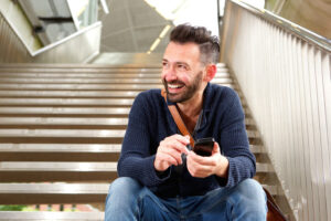 Smily guy with crinkled eyes holding his phone and sitting on staircase